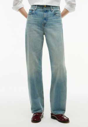 Personne portant un jean droit bleu clair, une chemise blanche à manches longues rentrée et des mocassins rouge foncé, debout, les mains dans le dos.