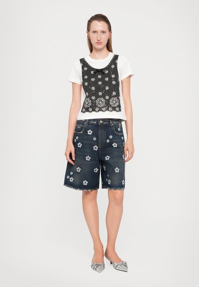 Vrouw in een wit T-shirt met een zwart bloemenkant overlay, donkerdenim shorts met witte bloemenprints en zilveren puntige lage hakken.