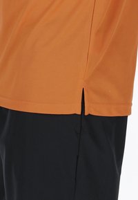 Orange kortærmet skjorte lavet af åndbart stof, har et sideslids og er parret med sorte shorts, hvilket fremhæver de kontrasterende farver.