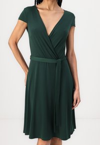 Robe portefeuille verte en tissu doux, dotée d'un décolleté en V profond, de manches courtes et d'une ceinture en tissu à la taille. Se transforme en jupe évasée.