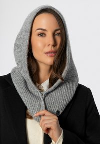 Sciarpa grigia a coste in maglia con cappuccio, caratterizzata da una consistenza liscia e chiusura con bottoni, indossata sopra una camicia bianca e un cappotto nero.