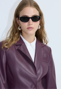 Blazer en cuir bordeaux avec un col cranté, fermeture à un seul bouton et détails de couture subtils, associé à une chemise blanche à col et des lunettes de soleil.