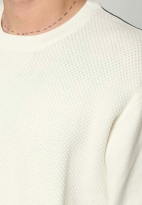 Witte gebreide trui met een structuur en een breipatroon, voorzien van een ribbelhalslijn en zwarte details langs de schoudernaden.