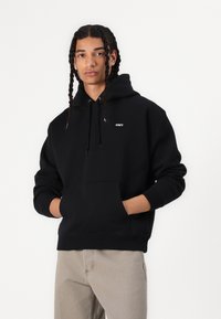 Schwarzer Baumwoll-Hoodie mit einer Vorderentasche, Kordelzugkapuze und kleinem weißen "OBEY"-Logo auf der Brust. Modell trägt graue Hose.