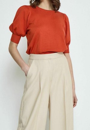 Femme portant un chemisier orange à manches courtes bouffantes et un pantalon beige large taille haute, une main dans la poche.