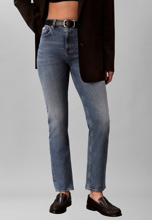 Vrouw draagt hoge taille rechte blauwe jeans met een zwarte riem, zwart cropped top, zwarte blazer en zwarte loafers, staand tegen een grijze achtergrond.