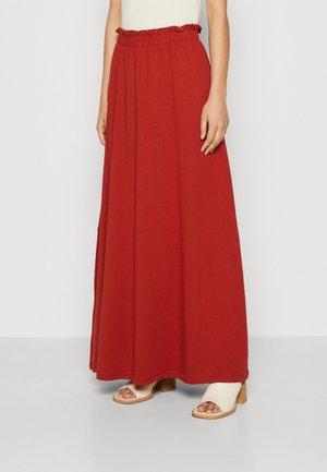 Vrouw draagt een lange, hoog opgesneden rode rok met een gerimpelde tailleband en opengewerkte crèmekleurige hakken, staand tegen een effen witte achtergrond.