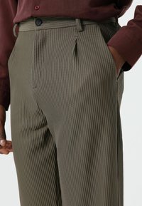 Olive green pleated trousers featuring vertical stripes, a single black button, and side pockets. Fabric appears textured and lightweight.