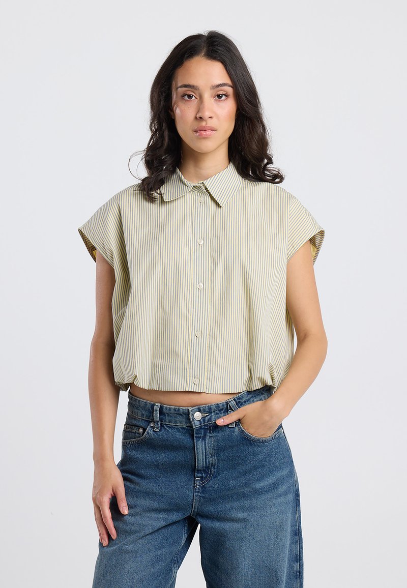 Cropped, short-sleeve shirt in light yellow with vertical stripes. Features a classic collar and button closure. Worn with high-waisted jeans.