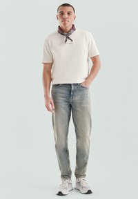 Man in een crèmekleurig overhemd met korte mouwen, vervaagde blauwe jeans, witte sneakers en een donkergekleurde sjaal om zijn nek, die staat met één hand in zijn zak.