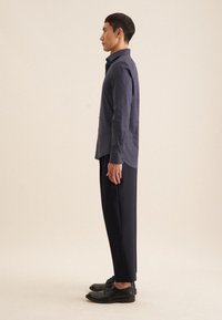 Marineblauwe polka dot blouse, lange mouwen, met knopen. Gecombineerd met zwarte tapered broeken en zwarte schoenen. Model staat in profielzicht.