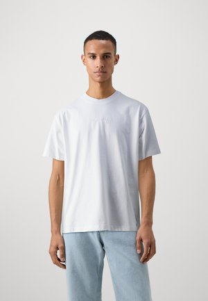 T-shirt en coton blanc à manches courtes, col rond et logo en relief subtil sur la poitrine. Porté avec un jean bleu clair.