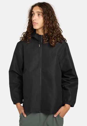 Junge Person mit langen, lockigen Haaren, die eine schwarze Kapuzenjacke mit Reißverschluss und grüne Hose trägt, die Hände in den Taschen und leicht nach links schauend.