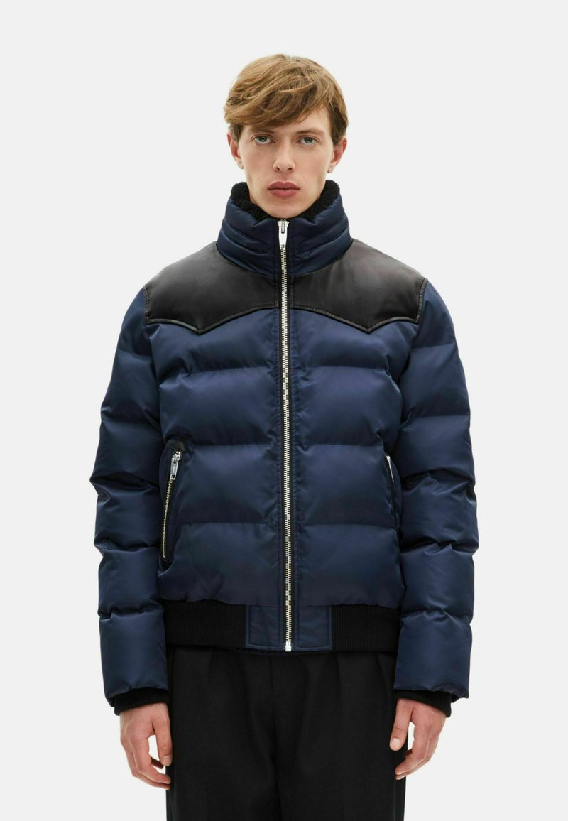 Marineblauer Steppmantel mit schwarzem Lederpasse, Reißverschluss, hohem Kragen und elastischen Bündchen. Mit Reißverschlusstaschen an den Seiten und gesteppter Struktur.