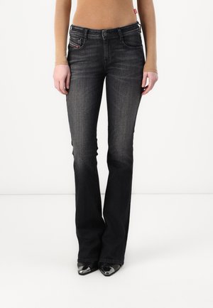 Kobieta ubrana w czarne, wyblakłe spodnie jeans typu bootcut z wysokim stanem oraz czarne buty z ostrym noskiem, stojąca na jednolitym białym tle.