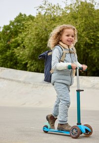 Blauer Roller mit zwei Rädern, strukturiertem Deck und robustem Lenker. Kind in grauem Stepp-Outfit fährt auf dem Gehweg und trägt einen blauen Rucksack.
