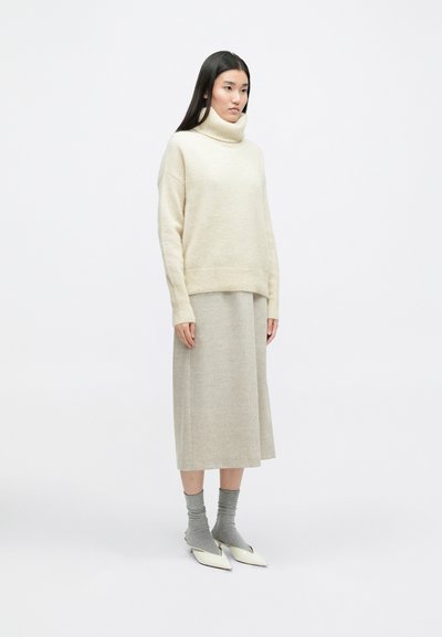 Μπεζ πουλόβερ με ψηλό λαιμό, μαλακή υφή, συνδυασμένο με μια ανοιχτό γκρι μίντι φούστα. Τα αξεσουάρ περιλαμβάνουν γκρι κάλτσες και λευκά παπούτσια με μυτερή μύτη.