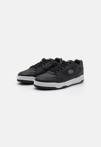 Fekete bőrsneaker perforált részletekkel, lapos fűzőkkel és fehér gumitalppal. Zöld logó díszítés található az oldalán.
