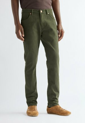Uomo che indossa pantaloni dritti verde oliva e scarpe stringate in suede marrone, in piedi su pavimento bianco.