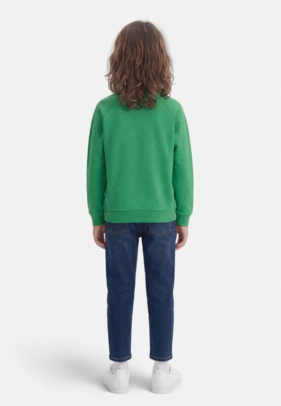 Παιδί με καστανά μαλλιά μήκους έως τον ώμο, που φοράει πράσινο φούτερ, μπλε τζιν, λευκές κάλτσες και λευκά αθλητικά παπούτσια, στέκεται γυρισμένο προς τα πίσω σε λευκό φόντο.