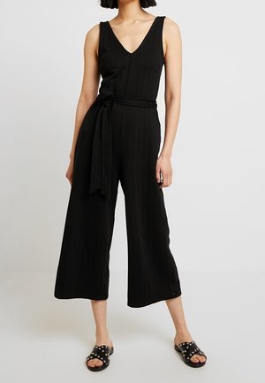 Kvinde iført ærmeløs, sort bredbenet jumpsuit med V-hals og bindebånd i taljen, kombineret med sorte slidssandaler med nitter.