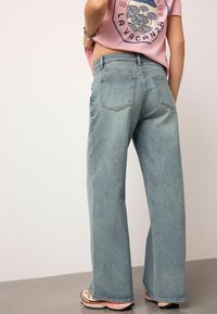 Lichtblauwe wijde jeans met een hoge taille, voor- en achterzakken en subtiele vervaging. Gecombineerd met een roze t-shirt met grafische print en sneakers.