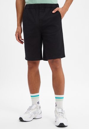 Uomo che indossa pantaloncini neri fino al ginocchio con fascia elastica in vita, calze sportive bianche con strisce blu e scarpe da corsa bianche.