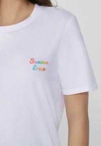Camiseta de algodón blanca con texto bordado colorido que dice "Amante del Verano" en varios tonos pastel. Cuello redondo y mangas cortas.