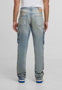 Personne portant un pantalon cargo en denim bleu clair avec des poches arrière et un débardeur blanc, se tenant sur un fond uni.