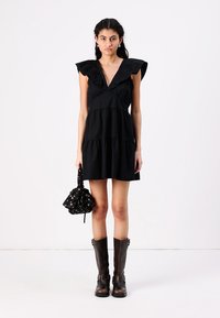 Vestido preto curto com design em camadas, mangas com franzido, decote em V profundo, combinado com uma mala de padrão floral e botas castanhas até ao joelho.