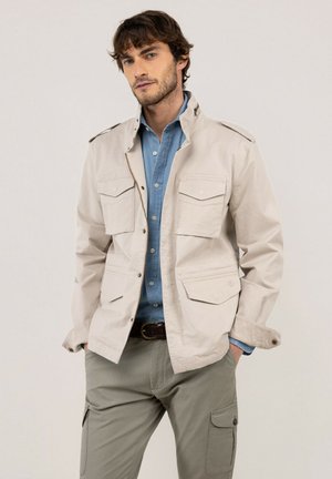 Hombre con chaqueta beige de trabajo sobre camisa azul con botones y pantalones cargo caqui con las manos en los bolsillos sobre fondo liso.