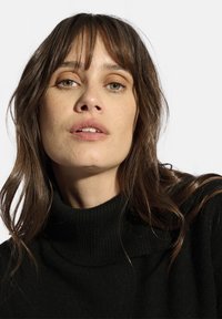 Schwarzer, gerippter Rollkragenpullover mit langen Ärmeln. Weiche Textur und lockere Passform. Das Model hat wellige braune Haare und ein neutrales Make-up.