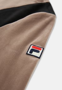Brun fleece stof med en sort stribe; har et broderet FILA-logo med røde og blå detaljer på en hvid baggrund.