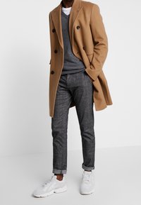 Personne portant un manteau double boutonnage camel, un pull gris en V, un pantalon gris à carreaux retroussé aux chevilles et des baskets blanches sur fond blanc.