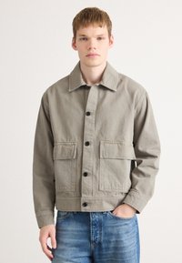 Grå bomullsjakke med knappelukking, to frontlommepåsydd, og krage. Brukt over blå jeans med en avslappet passform.