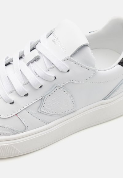 Baskets en cuir blanc avec lacets plats blancs, un logo latéral texturé, un col rembourré et une semelle en caoutchouc. Les accentuations incluent des détails gris et bleus.