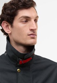 Casaco impermeável preto com gola alta, botões de pressão dourados e um detalhe em tecido vermelho no pescoço. Textura suave, design minimalista.