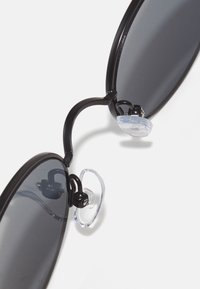 Black sunglasses with oval dark lenses, featuring a thin black frame, metal accents, and transparent nose pads for comfort and fit.