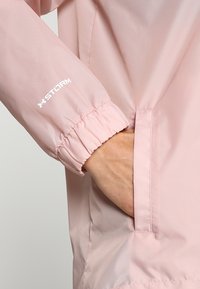 Chaqueta impermeable de color rosa claro con puños elásticos y un bolsillo lateral. Presenta un logo blanco de "STORM" en la manga. Textura suave y mate.