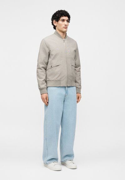 Jeune homme aux cheveux bouclés, debout, portant une veste beige zippée, un jean large bleu clair et des baskets blanches sur un fond uni.