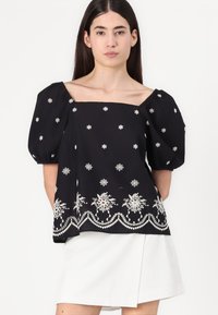 Blusa nera con maniche a sbuffo e scollo quadrato, con ricami floreali bianchi e dettaglio dell'orlo scallop.
