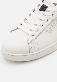Sneaker bianca in pelle con lacci bianchi piatti, suola in gomma testurizzata, logo argentato e accenti triangolari sui lati.