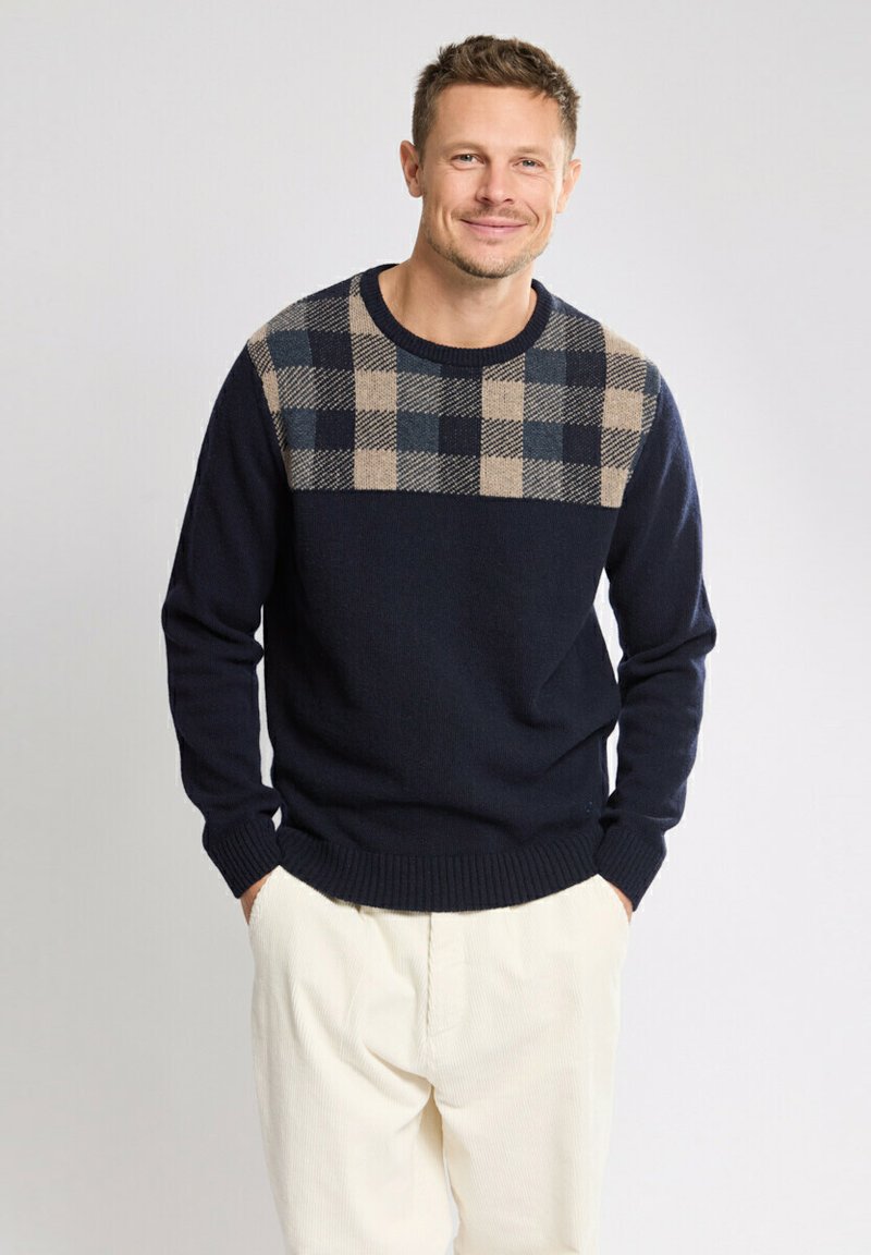 Homme souriant portant un pull bleu marine avec un motif à carreaux beige et bleu marine en haut, les mains dans les poches d'un pantalon blanc, sur fond uni.