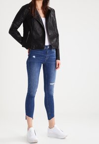 Giubbotto in pelle nera stile moto con tasche con zip, abbinato a jeans skinny blu usurati e sneakers bianche con zeppa.