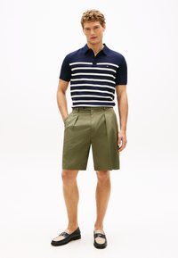 Marineblå polo med hvide striber, kombineret med olivengrønne plisserede shorts. Modellen bærer sorte og beige loafers.