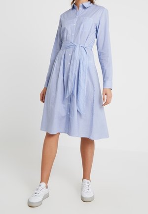 Robe chemise rayée bleu clair avec manches longues, boutonnée sur le devant, ceinture à nouer à la taille et une poche poitrine unique. Portée avec des baskets blanches.