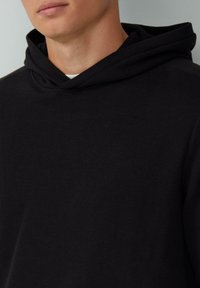 Schwarzer Kapuzenpullover mit gerippter Struktur, ausgestattet mit einer Kordelzugkapuze und lockerem Schnitt. Nahaufnahme zeigt die Stoffdetails.