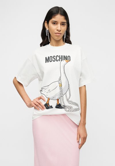 Λευκό βαμβακερό φαρδύ t-shirt με έγχρωμο γραφικό πάπιας που φοράει μαύρα παπούτσια και πέρλες, με το κείμενο "MOSCHINO" πάνω από το σχέδιο.