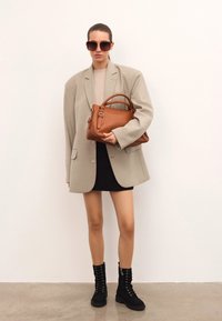 Kvinna som bär en överdimensionerad beige blazer, svart mini kjol, svarta snörstövlar och solglasögon, som håller en stor brun läderhandväska.
