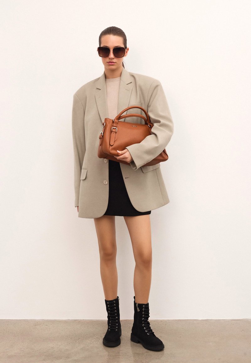 Kvinna som bär en överdimensionerad beige blazer, svart mini kjol, svarta snörstövlar och solglasögon, som håller en stor brun läderhandväska.